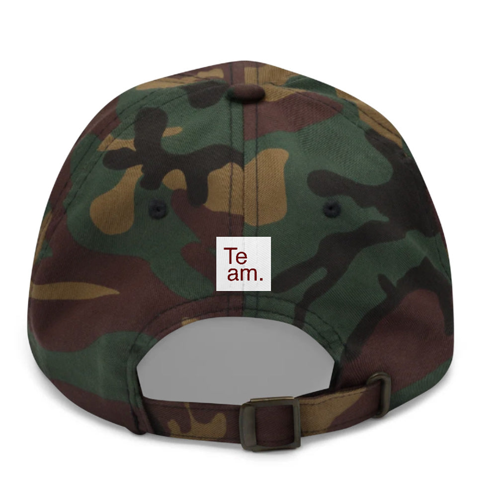 Rear view of a camouflage baseball cap with a small white square patch reading 'Te am.' on the back, shown on a plain white background.